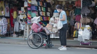 Bangkok, Tayland 'daki kalabalık Chatuchak Caddesi Marketi' nde tekerlekli sandalyedeki engelli bir kadın bir kadına ürün satıyor. - 4K Yatay Görüntü