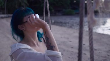 Sahilde salıncakta oturan havalı mavi saçlı kadının güneş gözlüğünü taktığı 4K yatay video.