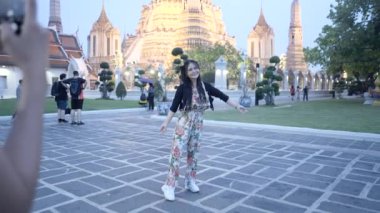 Asyalı kadın, Bangkok 'taki Budist tapınağının önünde farklı pozlar vermeye çalışıyor. Arkadaşı onu fotoğraflamak için akıllı ev tutuyor - 4K Yatay Video