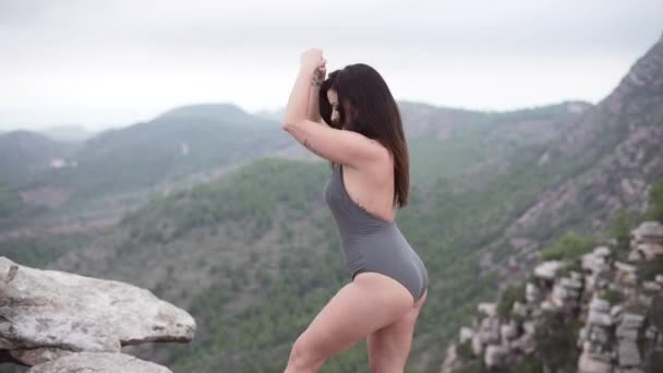 崖の上に立ってヨガの動きをする巨乳の女性 山の自然風景 水平ビデオ — ストック動画