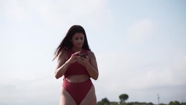 白人女性は屋外でスマートフォンを閲覧します 女性は曇りの背景水着を着て立っている 横ビデオ — ストック動画
