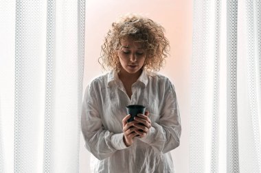 Kıvırcık saçlı, beyaz gömlekli, elinde bir fincan sıcak içecek olan genç bir kadın pencerenin yanında beyaz perdeli beyaz bir perdeyle dikilirken aşağıya bakıyor.