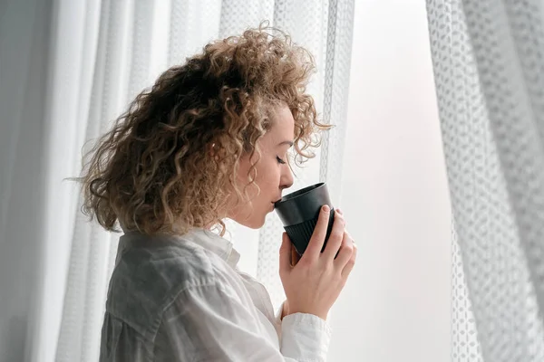 Side view of romantic lady with curly hair wearing white shirt standing near window with white curtain sipping coffee from black cup