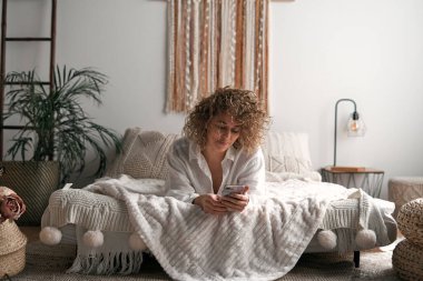 Beyaz tişörtlü, kıvırcık sarı saçlı, rahat bir yatakta yumuşak battaniye ve yastıklarla yatarken cep telefonunda gülümseyen ve mesajlaşan pozitif genç bir kadın.