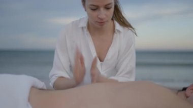 Tanınmayan adam sahilde yağ ile sırt masajı yaptırıyor. Güçlü elleri olan genç beyaz kadın masaj yapıyor - 4K yatay video