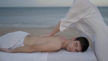 Plajda tedavi masajı yapan beyaz, ikili olmayan genç bir insan. Masör sırt masajına baskı uyguluyor - 4K yatay video