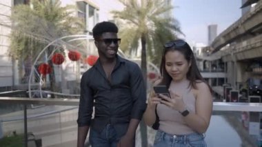 Asyalı ve Afrikalı arkadaşlar Bangkok alışveriş merkezinin dışında etkileşime girdiler, kız 4K yatay videosunu işaret ediyor.