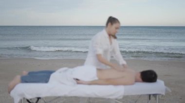 Adam plajdaki masaj masasında dikkatsiz bir şekilde yatıyor. Arka planda hareketli deniz var. 4K yatay video.