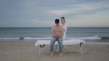 İkili olmayan müvekkil dışarıda terapi sırasında masaj masasına oturur. Kadın masöz sırt masajı yapıyor - 4K Yatay video