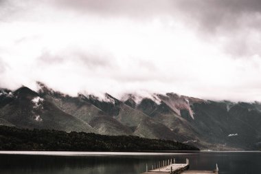 Çok sakin bir göl manzarası ve dağların saflığıyla çevrili yalnız eski bir iskele. Gökyüzünün gri bulutlarının gölgeleri suya, Nelson göllerine, yeni göllere yansıyor.