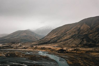 Güneşsiz bir günde bulutlar ve sisle kasvetli bir gökyüzü altında yüksek ağaçsız ve karlı dağları aşan yol kenarındaki yavaş akan nehir ve büyük ovanın havadan görünüşü.
