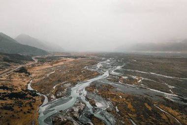 Nehir, bulutlu gri gökyüzünün altında, dağların kıyısında uzanan bir yol ile büyük kahverengi bir ovada kıvrılır, aşçı, Yeni Zelanda - seyahat kavramı