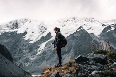 Boynunda SlR kamerasıyla genç beyaz bir adam kalın bir ceket ve spor ayakkabılar sakin ve rahat bir şekilde göl kenarındaki güzel manzarayı ve karlı dağları seyrediyor.