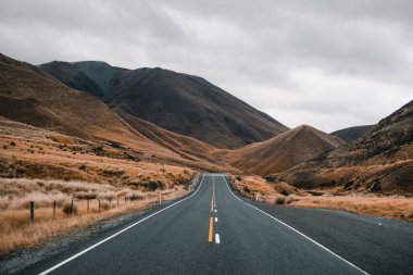 Trafiksiz uzun bir yol ıssız ağaçsız bir arazide ıssız yüksek dağların arasında kayboluyor bir sürü gri bulutlu, dağ aşçısı, Yeni Zelanda - seyahat konsepti