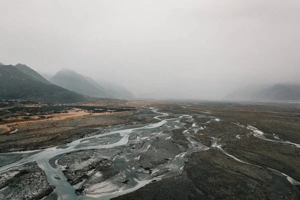 sad and desolate landscape with a poor river in the calm and silent plain that crosses the mountains and is lost in the fog under a very cloudy sky near the sea, mount cook, new zealand - Travel