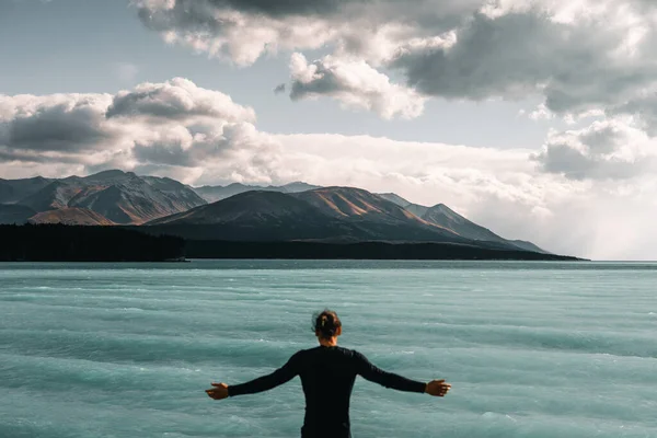 caucasian boy with long messy bun hair standing on his back with open arms and a black t-shirt looking at the wild sea and mountains under a stormy sky, mount cook, new zealand - Travel concept