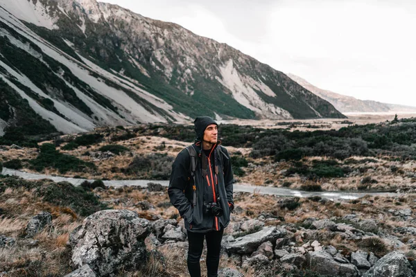 happy and calm young caucasian man enjoying the landscape with a black hoodie a camera hanging and hands in pockets on a very cold day under a gray sky near the snowy mountains and on the rocks, mount