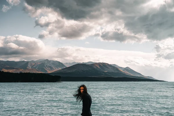 young caucasian man with long disheveled hair wearing t-shirt and black pants looking at camera from rough sea by mountains under stormy sky with gray and black clouds, mount cook, new zealand -