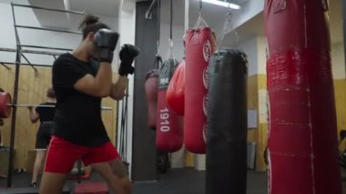 Spor salonunda boks tekniği uygulayan güçlü bir atlet. Eldiven takarak kum torbasına vuran adam - 4K yatay video
