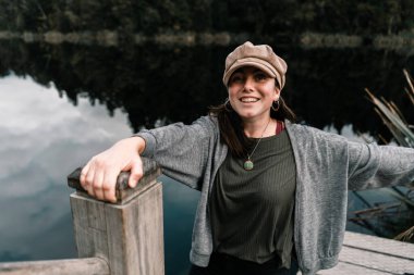 Kahverengi şapkalı, gri ceketli, siyah pantolonlu, dans eden, yeşil ormandaki sakin ve sessiz soğuk su gölünün kıyısındaki ahşap çardakta mutlu mesut dans eden beyaz bir kız.