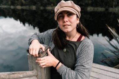 Kafkasyalı genç bir kız güzel kahverengi şapka gri ceket ve yeşil tişört giyiyor sevimli yuvarlak kolye kameraya bakıyor sakin gölün iskelede büyük ahşap korkuluklara yaslanıyor, Yeni Zelanda buzulları -