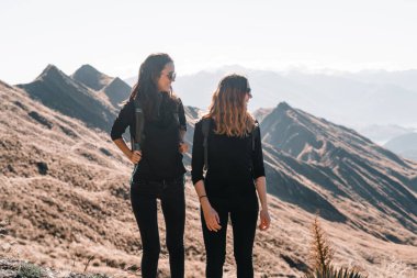 Siyah tişörtler ve pantolonlar içinde iki beyaz kız, sessiz ve yalnız bir dağın zirvesinde manzaranın güzelliğini izlerken mutlu bir şekilde gülüyorlar.