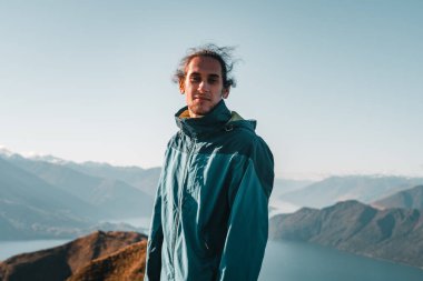 Dağınık, mavi ceketli beyaz çocuk kameraya bakıyor sakin ve sessiz bir dağ zirvesinde gülümsüyor büyük mavi su gölü üzerinde, Roys Peak, Yeni Zelanda - seyahat konsepti