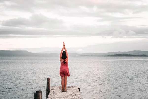 Caucasian young woman wearing black bracelet pink dress and sandals standing with feet together and arms outstretched upwards at end of wooden pier under cloudy sky in Okere, new zealand - Nature