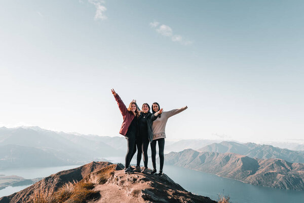 three caucasian girls in black pants looking at camera with arms outstretched smiling and happy having fun having fun hugging from top of mountain enjoying nature, roys peak, new zealand - Travel