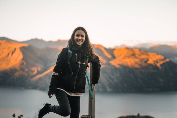 caucasian cute brunette girl in black jacket black pants and sneakers holding onto wooden stick with knee raised and standing on one leg happy and content on mountain summit at sunset, roys peak, new