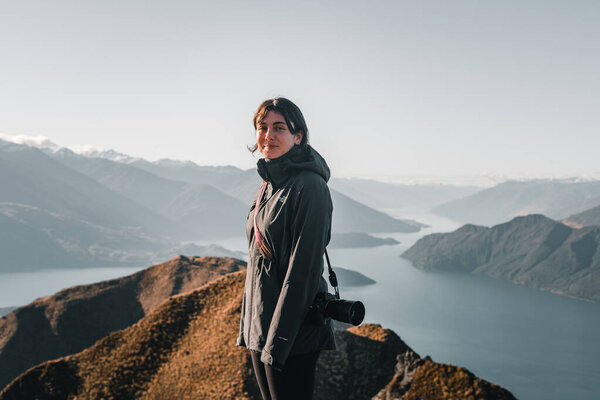 blue-eyed caucasian girl wearing black pants coat and reflex camera hanging from left shoulder smiling and enjoying great lake scenery from mountain summit, roys peak, new zealand - Travel concept