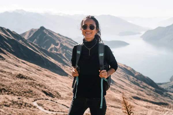 disheveled caucasian young woman with big earrings in her ears black t-shirt and black pants necklace on her neck black backpack smiling happy and content on the mountain summit overlooking the big