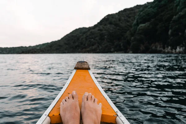 feet together resting on the prow of a white and orange wooden canoe heading into the forest navigating the lake waters at sunset, tarawera lake, new zealand - Lifestyle travel concept