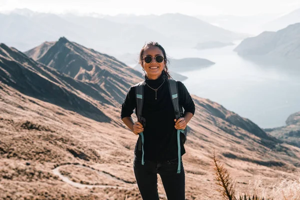 standing caucasian young woman in sunglasses looking at camera with gray backpack black t-shirt and black pants smiling from mountain summit near big blue lake and stunning mountains, roys peak, new