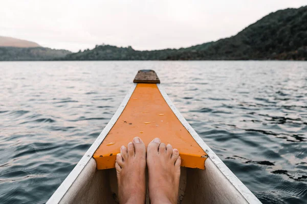 feet together inside an orange and white wooden canoe sailing towards the mountains of the lakeside island under a gray sunset sky, tarawera lake, new zealand - Lifestyle travel concept