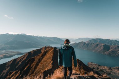 Mavi ceketli ve siyah pantolonlu beyaz bir çocuk dağın tepesindeki bir taşa tırmanıyor. Büyük mavi gölü çevreleyen etkileyici dağların güzel manzarasının tadını çıkarmak için.