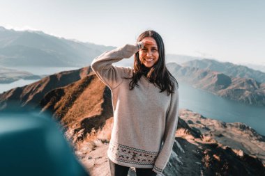 Esmer, beyaz tenli, kahverengi gözlü, uzun saçlı, gri kazaklı, sağ eli yüzünü kaplayan siyah pantolonlu, doğanın ortasında, sakin bir manzarada dağın tepesinden gülümseyen bir kız.