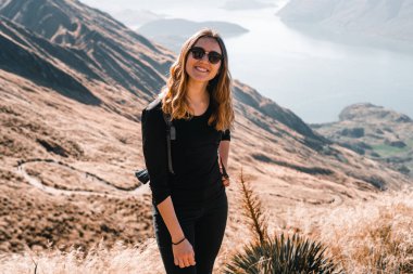 Sırtında güneş gözlüğü ve sırt çantasıyla beyaz saçlı genç bir kadın kameraya gülümsüyor. Sakin ve sakin bir dağın zirvesinde, büyük mavi gölün yanında, Roys Peak, Yeni Zelanda -