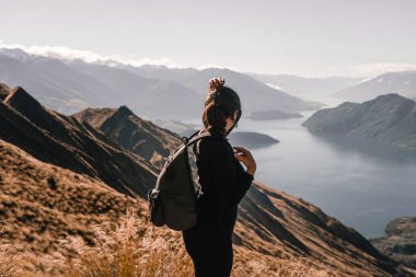 Sırtında sırt çantası olan beyaz, siyah pantolonlu genç bir kadın. Sol eli başının üstünde. O sakin ve rahat bir şekilde büyük mavi gölün manzarasına ve etkileyici manzarasına hayran.