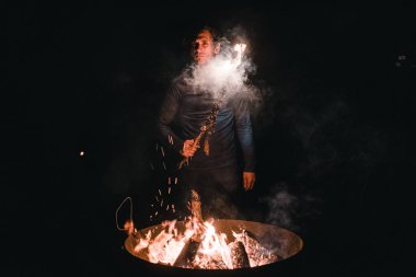 Büyük metal bir konteynırın içinde yanan şenlik ateşi ve siyah tişörtlü ve siyah pantolonlu beyaz çocuk kameraya ciddi ciddi bakıyor ve geceleri sağ eliyle yanan kuru bir dal tutuyor.