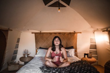 Rahat, mutlu, sakin, beyaz bir kadın. Yatağında bağdaş kurmuş, elinde bir fincan kahveyle kamp çadırında oturuyor. Hayat tarzı seyahat kavramı.