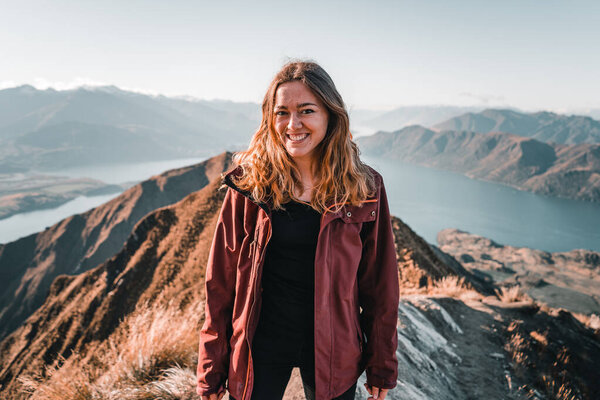 blonde caucasian young girl wearing brown jacket black t-shirt and black pants happy and content smiling having fun from the top of the mountain in wonderful beautiful setting in nature, roys peak