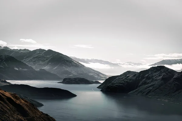 nice landscape of calm blue water of the big lake and the impressive dark rocky mountains without vegetation in a calm and sad environment under a slightly cloudy sky, roys peak, new zealand - Travel