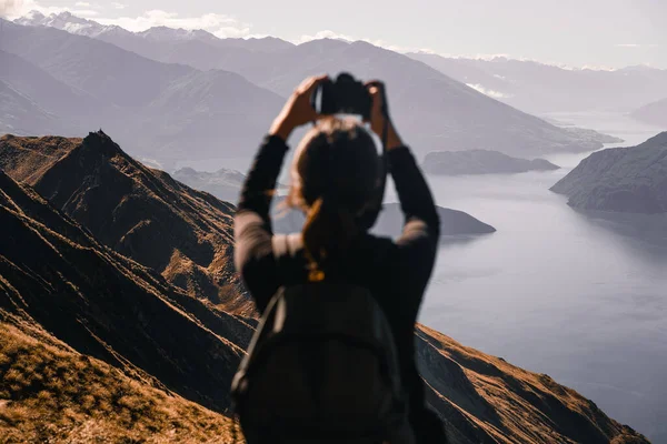 caucasian girl with backpack on her back black t-shirt and hair in a ponytail taking a photo with reflex camera with arms outstretched upwards on mountain summit near big lake and stunning mountains
