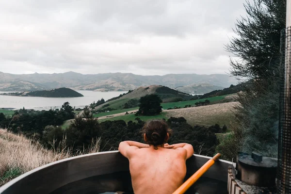 Caucasian young man without clothes on his back taking a bath and enjoying the nice scenery inside a metallic hot water jacuzzi relaxing in the mountains far from civilization near the forest trees