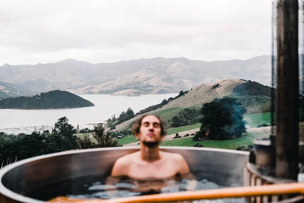Caucasian boy with long collected hair relaxed and calm with closed eyes inside a small round wooden jacuzzi and hot water in the middle of the forest near the calm trees and greenery in the middle of