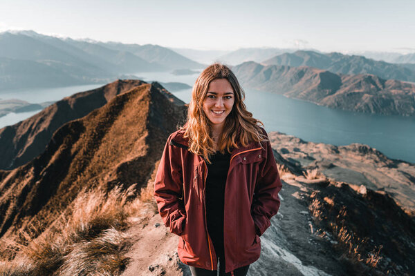 blonde caucasian young woman with brown eyes with hands in pockets of brown jacket black t-shirt and black pullover smiling happily at camera from a mountain summit calm and relaxed enjoying the