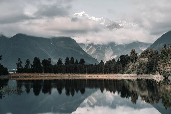 calm and lonely landscape of snowy mountains covered by clouds stunning stormy day at calm water lake amid nature vegetation and green forest, new zealand glaciers - Nature concept