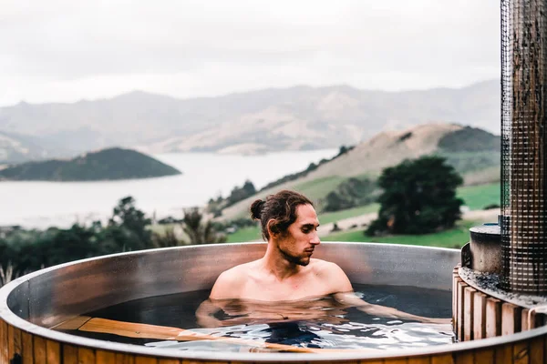 caucasian guy with long collected hair with a beard serious relaxed and calm inside a small round wooden jacuzzi and hot water in the middle of the forest near the calm trees and vegetation in the