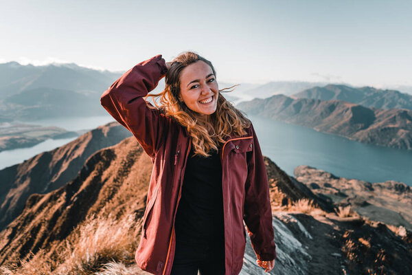 blonde caucasian girl wearing nice black t-shirt jacket smiling at camera with right hand on head happy and content from top of mountain with nice scenery of big blue lake and rocky mountains, roys